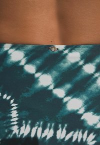 Badtyg i teal och vitt med batikmönster, har en slät textur och pryds av en liten metallisk detalj vid midjan.