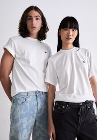 Dva modelky v bílých tričkách Lacoste a stylových džínách, stojící vedle sebe na jednobarevném pozadí.
