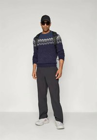 Marineblaue Strickjacke mit geometrischen Mustern, dunkle Hosen, weiße Turnschuhe, Rucksack, Kappe und Sonnenbrille. Lässige Outdoor-Bekleidung.