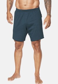 Mann in dunkelblauen elastischen Shorts, barfuß stehend, mit sichtbar tätowierten Armen vor einem weißen Hintergrund.