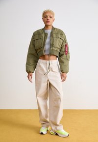 Avskuren olivgrön bomberjacka med två framfickor, matchad med krämfärgade vida byxor och sneakers med neondetaljer. Slät textur.