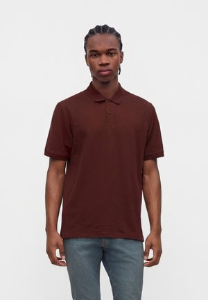 Jeune homme aux cheveux tressés portant un polo marron à manches courtes et un jean bleu, debout devant un fond blanc uni.