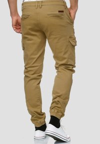 Khaki színű, elasztikus mandzsettaszárral rendelkező jogger nadrág, oldalsó cargo zsebekkel és hátsó welt zsebekkel. Simaszövet, enyhe fényű. Fehér sportcipő fekete kiegészítőkkel.