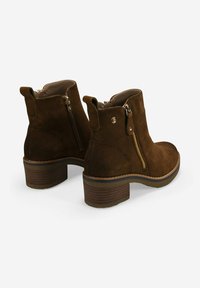 Botas de ante marrón hasta el tobillo con cremalleras laterales, tiradores y tacones bloque apilados sobre un fondo blanco.