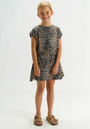 Smiling girl standing with hands held loosely, wearing a short-sleeve leopard print dress and tan sandals against a plain background.