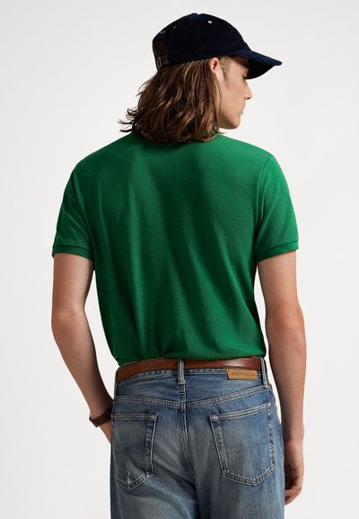 Πράσινο βαμβακερό πόλο μπλουζάκι με κοντά μανίκια, συνδυασμένο με ξεθωριασμένο μπλε τζιν και μπλε σκούφο, φαίνονται από πίσω. Ορατή καφέ δερμάτινη ζώνη.