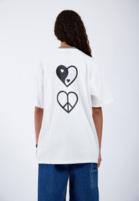 Camiseta blanca de algodón con un diseño gráfico negro que presenta dos corazones, uno que muestra un patrón de yin-yang y el otro un símbolo de paz.