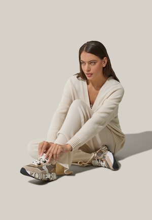Sneaker beige con lacci, dotate di una parte superiore testurizzata e multicolore con accenti mimetici e lettering audace sul lato, posizionate su uno sfondo neutro.