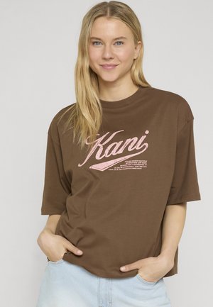 Blondynka z długimi włosami, ubrana w luźny brązowy T-shirt z różowym napisem "Kani", ręce w kieszeniach, uśmiechnięta, stojąca na jednolitym tle.