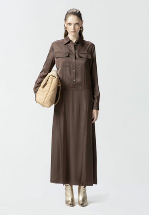 Abito lungo in satin marrone con due tasche sul petto e bottoni, abbinato a una borsa trapuntata beige e scarpe con punta dorata.