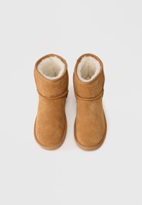 Botas de tornozelos em camurça bege com um forro de pelúcia branca macia, apresentando um bico arredondado e detalhes costurados no exterior.