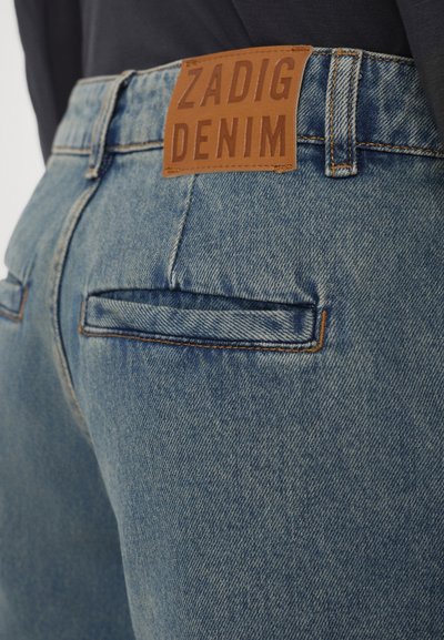 Jeans de mezclilla azul con una etiqueta de cuero que dice "ZADIG DENIM". La tela tiene una trama texturizada y costuras visibles.