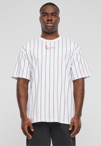 Mand iført en hvid T-shirt med korte ærmer, der har fine sorte lodrette striber og et lille rødt signaturdetalje centralt på brystet, kombineret med sorte bukser.