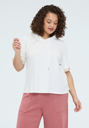 Frau mit lockigem Haar, die einen weißen Kurzarm-Hoodie und rosa Hosen trägt, hält eine Kapuzenkordel vor einem schlichten hellen Hintergrund.