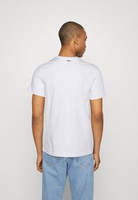 Camiseta de algodón blanca con mangas cortas, cuello redondo y una etiqueta de marca pequeña en la parte posterior. Combinada con unos pantalones vaqueros de denim azul claro.