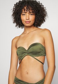Olivengrønn bandeau-bikinitopp med rynkedesign, myk tekstur og kryssede stropper. Har en strukturert form og glatt overflate.