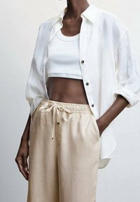 Witte linnen overhemd met knopen over een witte crop top, gecombineerd met beige trekbroek met een relaxte pasvorm en een lichte stof.