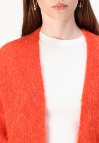 Oransje fluffy cardigan med åpen front, lagd over en hvit ribbestrikket t-skjorte. Den teksturerte stoffer er myk og varm.
