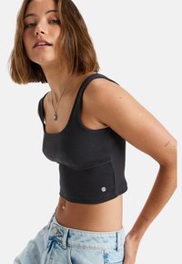 Čierny rebrovaný crop top s širokými ramienkami, priliehavým strihom a detailom vrecka na boku. Spárovaný s svetlo modrými džínsovými šortkami.