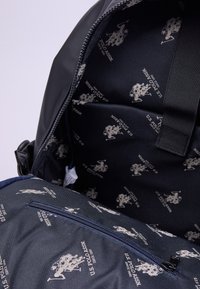 L'interno dello zaino nero presenta un tessuto blu navy foderato con ripetuti loghi grigi della U.S. Polo Assn., evidenziando una superficie liscia e una tasca con cerniera.