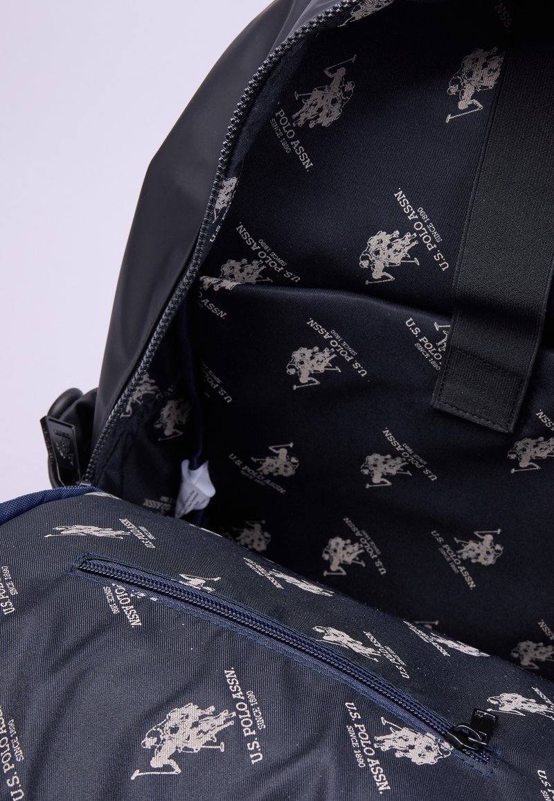 L'interno dello zaino nero presenta un tessuto blu navy foderato con ripetuti loghi grigi della U.S. Polo Assn., evidenziando una superficie liscia e una tasca con cerniera.