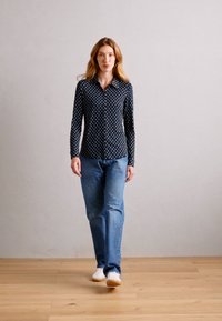 Marineblå skjorte med hvite prikker, kombinert med lyseblå jeans og hvite joggesko, satt mot en nøytral bakgrunn.