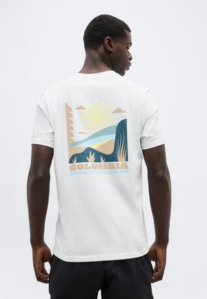 Hombre con camiseta blanca con un gráfico colorido de sol y montaña y texto "Columbia Sportswear Co." en la parte posterior, de espaldas.