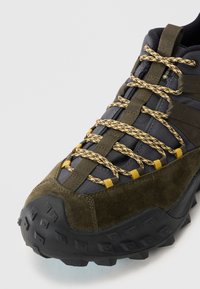 Chaussure robuste noir et vert foncé avec une semelle en caoutchouc texturée, des lacets jaunes et blancs, et un mélange de matériau en daim et en tissu.