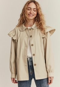 Beige jakke med flæset ærmer og en dekorativ krave. Har store knapper, blød stof og en afslappet silhuet.