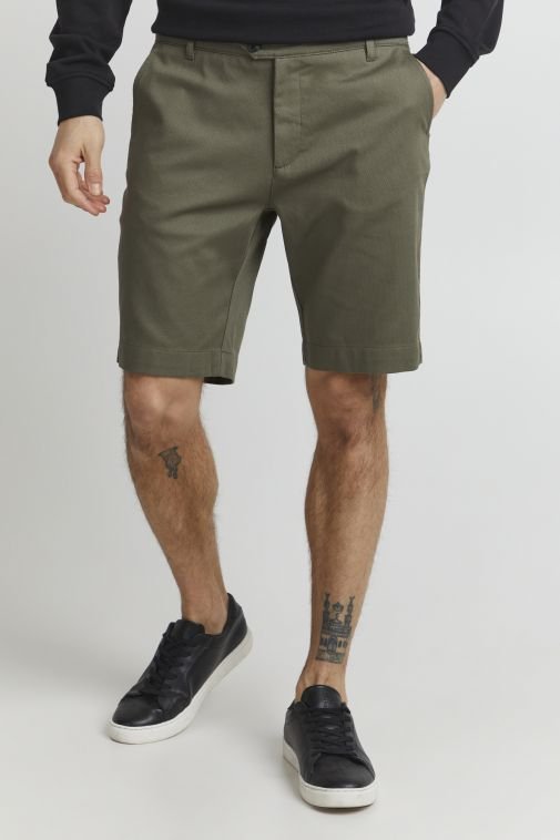 Shorts en coton vert olive avec une coupe plate, deux poches latérales et un pli subtil. Association avec des baskets noires, mettant en avant un look décontracté.