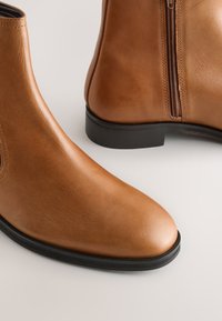 Bottes courtes en cuir marron à texture lisse, bout arrondi et semelles en caoutchouc noires ; dotées d'une fermeture éclair latérale pour un enfilage facile.