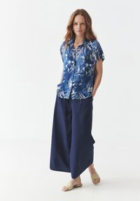 Camicia a maniche corte blu con motivo floreale e vestibilità rilassata, abbinata a pantaloni a gamba larga blu navy e sandali beige a scivolo.
