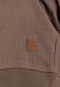 Tejido acanalado marrón con una superficie texturizada y un parche de cuero rectangular que presenta el texto "KEY LARGO" y un logo gráfico.