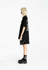 Persona con cabello rubio claro lleva una camiseta negra de gran tamaño con un gráfico blanco en la espalda, pantalones cortos negros con detalle de cadena, calcetines negros y zapatos negros con textura.