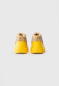 O pereche de adidași UGG bej și galbeni văzuți din spate, având închideri cu șnur la gleznă și tălpi groase galbene.