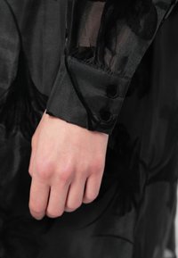 Χέρι με ανοιχτόχρωμο δέρμα που ξεκουράζεται κοντά στον καρπό, φορώντας μανίκι από μαύρο διάφανο ύφασμα με σατέν μανσέτα και δύο μαύρους κούμπες.