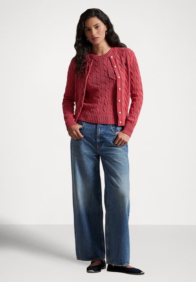 Sarkans adīts kardigans ar baltiem pogām pār saskaņotu džemperi, kombinēts ar augsta vidukļa platajiem džinsa biksēm un tumši ziliem slip-on apaviem.