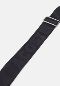 Cinturino in tessuto blu navy con testo "LACOSTE" impresso, arricchito da una fibbia metallica argentata. La superficie testurizzata aggiunge profondità al design.