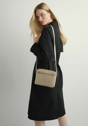 Blondynka w czarnym płaszczu trencz trzyma beżową torbę na ramię z zamykanymi na suwak kieszeniami na jednolitym jasnym tle.