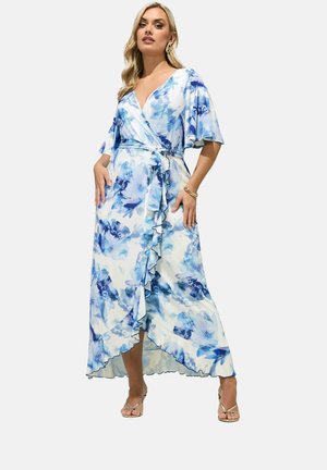 Vrouw die een blauwe wikkeljurk met bloemenprint draagt, voorzien van vlindermouwen en een gerimpelde zoom, gecombineerd met gouden sandalen met bandjes, tegen een effen achtergrond.