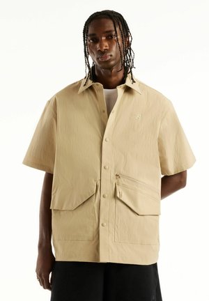Jeune homme aux cheveux tressés portant une veste beige à manches courtes avec de grandes poches et un pantalon noir, debout devant un fond blanc.