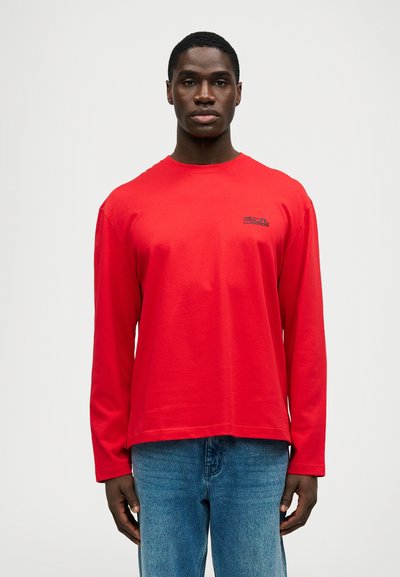 Νεαρός άνδρας φοράει ένα κόκκινο μακρυμάνικο πουκάμισο με μικρό μαύρο κείμενο στο στήθος και μπλε τζιν, στέκεται μπροστά σε μια απλή λευκή εικόνα.