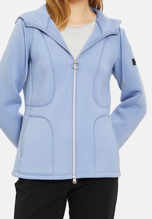 Frau trägt einen hellblauen Kapuzenpullover mit Reißverschluss über einem grauen Shirt und schwarzen Hosen, mit den Händen in den Kapuzenpullover-Taschen.