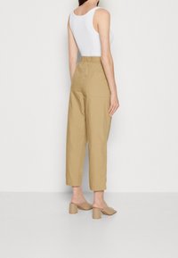 Donna in piedi contro un muro bianco, indossando un top senza maniche bianco, pantaloni beige a vita alta e cropped, e mule beige con tacco a blocco.