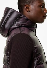 Gilet imbottito di colore viola scuro con un esterno liscio e lucido e maniche in tessuto nero a coste. Presenta un cappuccio e una chiusura con zip.