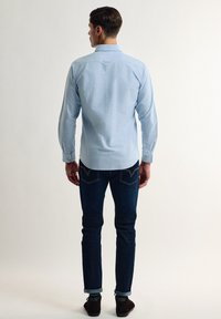 Camisa de manga larga azul claro con puños de botón, combinada con vaqueros oscuros. La camisa presenta un dobladillo curvado y pliegues en la parte posterior para un mejor ajuste.