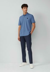 Hellblaues Polohemd mit einem schwarzen Logo-Patch, kombiniert mit dunklen Hosen und weißen Sneakers, vor einem hellen Hintergrund stehend.