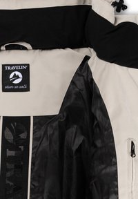 Beige travel jacket met zwarte accenten, met een merklogo en een gladde, glanzende textuur. Inclusief ritszakken en een comfortabele kraag.
