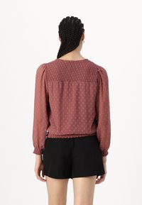 Blusa color marrone con punti texturizzati, corpino arricciato e maniche lunghe a sbuffo. Orli sfilacciati in fondo e alle estremità delle maniche. Indossata con pantaloni corti neri.