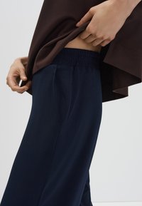 Marineblauwe sportbroeken met een elastische tailleband en zijzakken, gecombineerd met een bruin losvallend shirt. De gladde stoftextuur is zichtbaar.